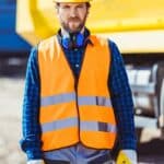 Worker in reflective vest and hardhat standing at construction site in front of big yellow truck and